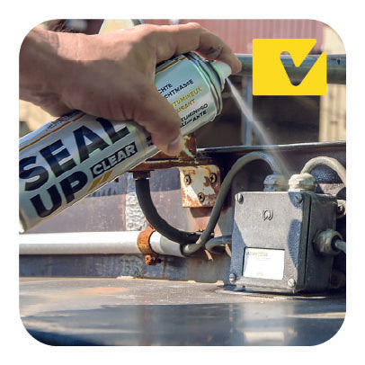 Un'immagine che mostra una mano mentre utilizza una bomboletta spray di sigillante trasparente con il marchio 'Seal Up Clear' su una connessione meccanica. La scena si svolge all'aperto, con un dispositivo metallico e tubi visibili.